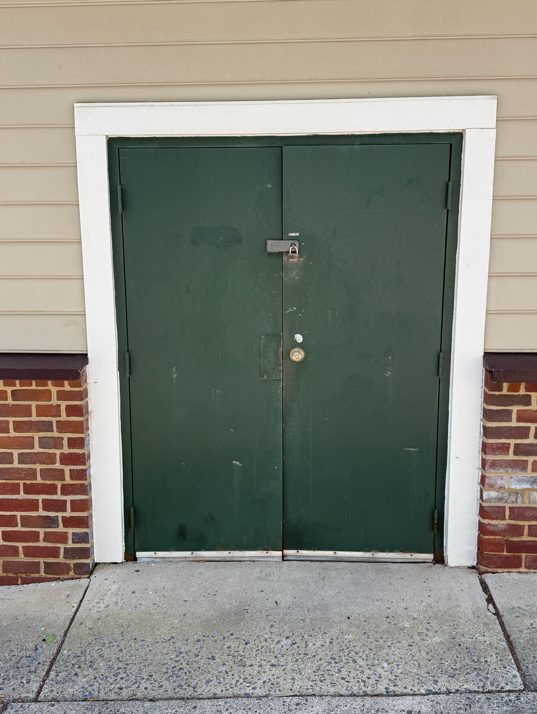 The original rear service door before replacement: a green painted steel double door with visible corrosion along the bottom edge and worn hardware, set in a white frame in a brick wall.