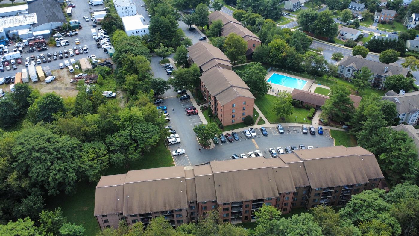 Aerial photograph of a multi-building garden apartment community, showing long shingled rooflines, surface parking and mature tree cover.