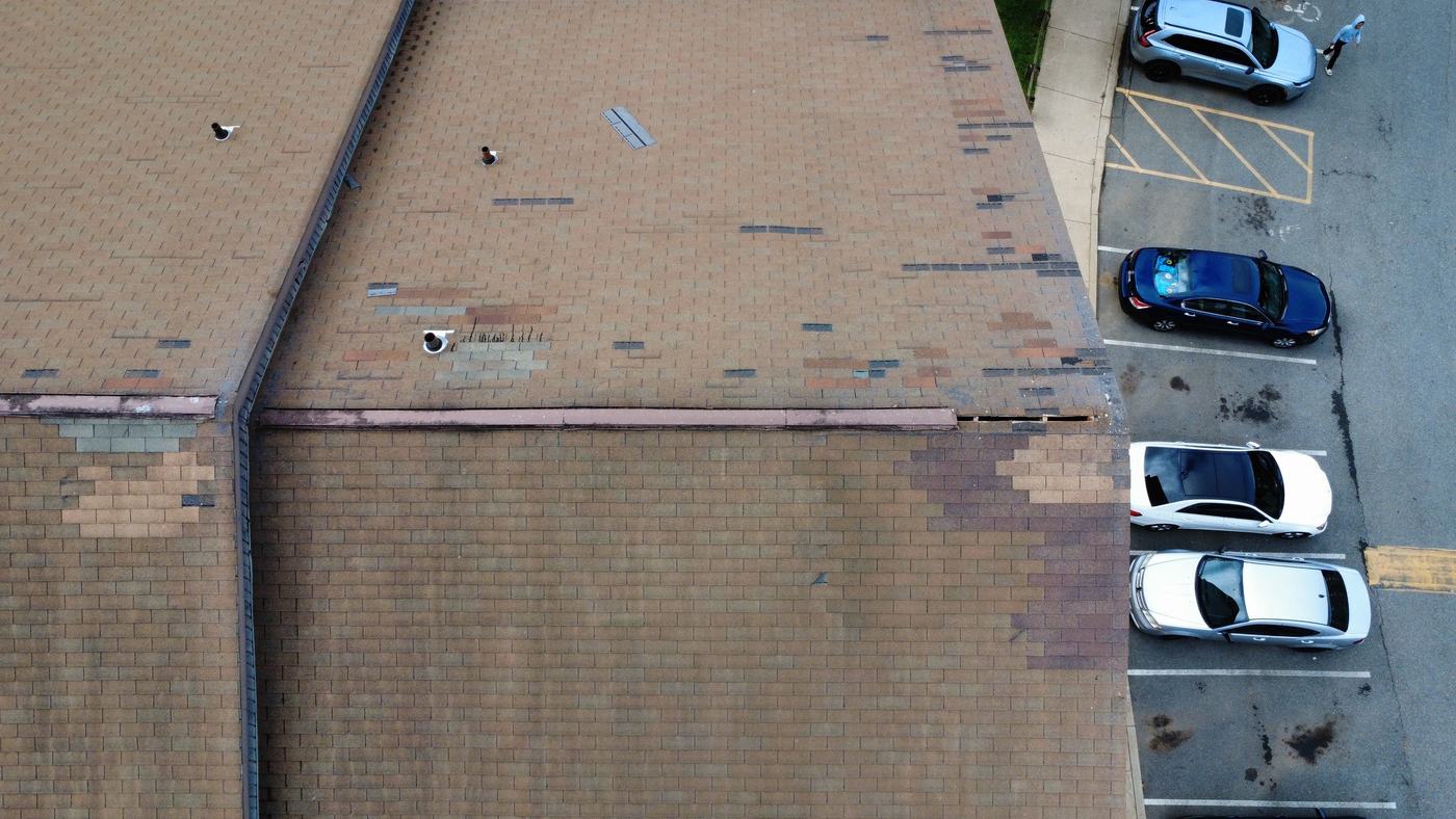 Overhead view of a shingle roof field above a parking lot, showing patches of mismatched shingles from earlier spot repairs and uneven granule wear.